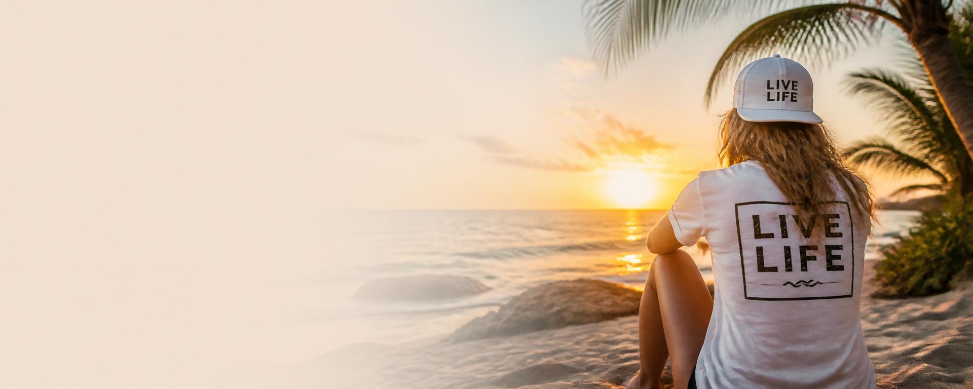 Woman wearing LIVE LIFE® apparel while sitting beside the ocean at sunset.